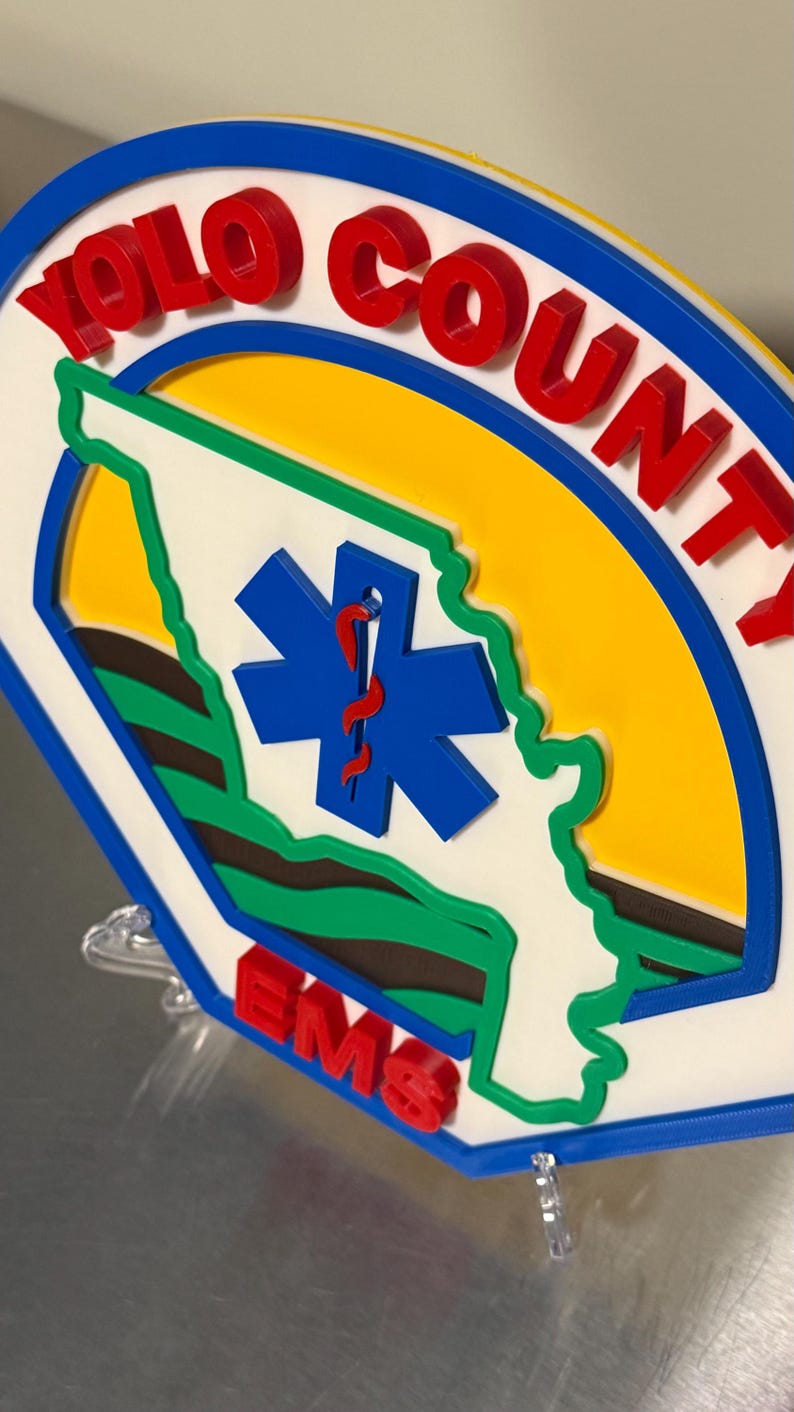 May include: A circular sign featuring the words "YOLO COUNTY" in red, with a blue border. The sign includes a blue star of life symbol and a green outline of a state. The letters "EMS" are in red.