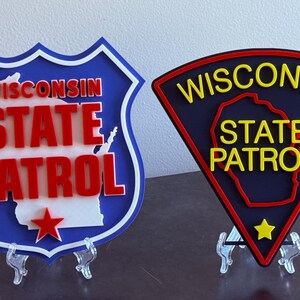 May include: Two Wisconsin State Patrol emblems. One is shield-shaped with a blue border, white interior, red text, and a red star. The other is a black triangle with yellow text, a yellow star, and a red border.