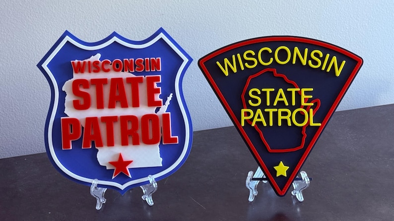 May include: Two Wisconsin State Patrol emblems. One is shield-shaped with a blue border, white interior, red text, and a red star. The other is a black triangle with yellow text, a yellow star, and a red border.