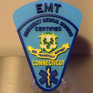 Op de afbeelding: Een lichtblauw EMT-badge met de woorden "EMT Emergency Medical Services Certified Connecticut" in wit en geel. De badge heeft een levensster-symbool en een staatszegel.