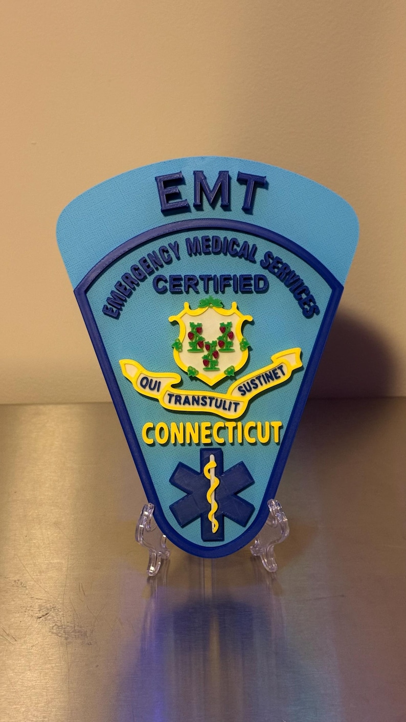 Op de afbeelding: Een lichtblauw EMT-badge met de woorden "EMT Emergency Medical Services Certified Connecticut" in wit en geel. De badge heeft een levensster-symbool en een staatszegel.