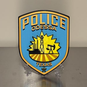 May include: A shield-shaped emblem with the words "POLICE ODESSA TEXAS" in blue and yellow. The emblem features a yellow oil derrick, factory, and star. The background is light blue.