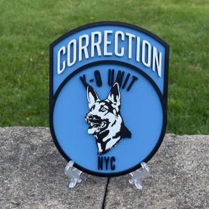 May include: A blue and black sign with the words "CORRECTION K-9 UNIT NYC" in white lettering. The sign features a black and white graphic of a German Shepherd dog.