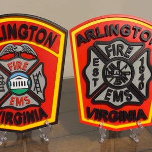May include: Two red and yellow Arlington, Virginia fire department plaques with black lettering and a black and gray fire department emblem. The plaque on the left has a bird with outstretched wings above the emblem. The plaque on the right has the words "EST 1940" on the emblem.