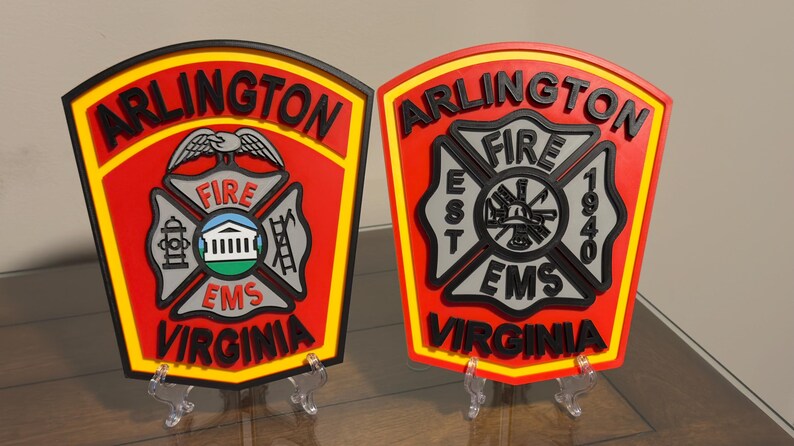 May include: Two red and yellow Arlington, Virginia fire department plaques with black lettering and a black and gray fire department emblem. The plaque on the left has a bird with outstretched wings above the emblem. The plaque on the right has the words "EST 1940" on the emblem.