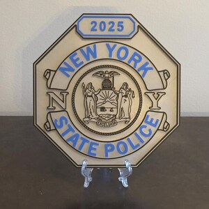 May include: A wooden plaque with a blue and black New York State Police badge design. The badge features the New York State seal and the text "NEW YORK STATE POLICE" in a scroll design. The year "2025" is displayed in a blue rectangle at the top of the plaque.