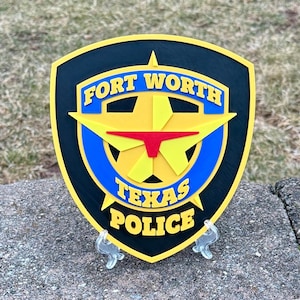 May include: A black and yellow police badge with a five-pointed star in the center. The badge reads "FORT WORTH TEXAS POLICE" in yellow letters.