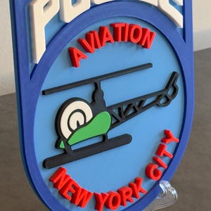 May include: A blue and white 3D printed sign with the words "POLICE AVIATION NEW YORK CITY" in red and white letters. A black helicopter with a green body is in the center of the sign.