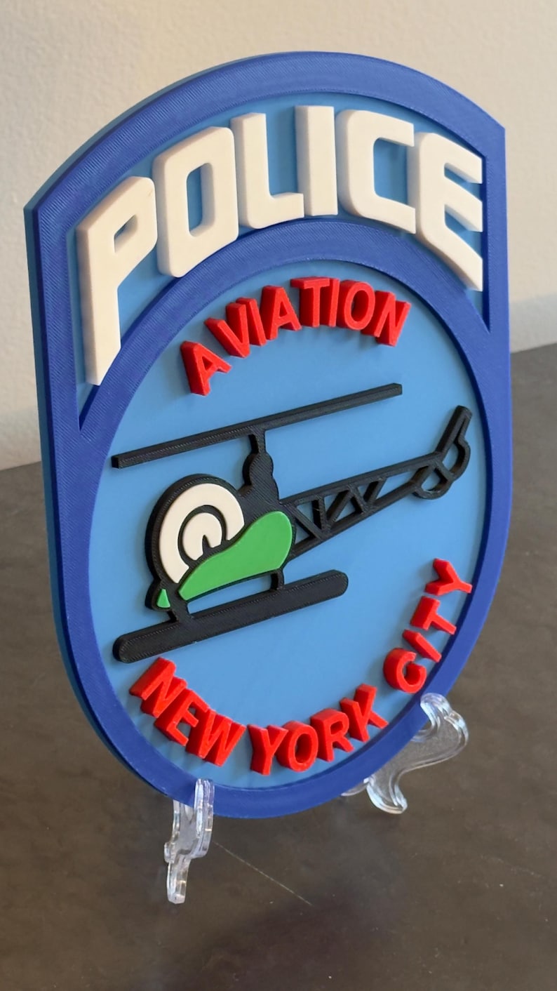 May include: A blue and white 3D printed sign with the words "POLICE AVIATION NEW YORK CITY" in red and white letters. A black helicopter with a green body is in the center of the sign.