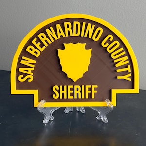 May include: A brown and yellow San Bernardino County Sheriff's badge with a shield in the center. The badge is mounted on a clear plastic stand.