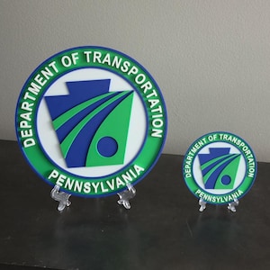 May include: Two circular signs featuring the Department of Transportation Pennsylvania logo. The design includes a blue keystone shape with green curved lines and a circle on a white background, surrounded by a green border with white text.