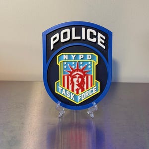 May include: A blue, black, and yellow shield-shaped wall plaque featuring the text POLICE at the top and TASK FORCE at the bottom. The center displays a graphic of the Statue of Liberty in red and white with the letters NYPD above it.