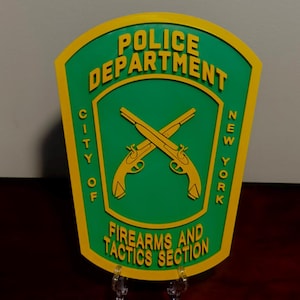 May include: A green and yellow Police Department emblem from New York City. The emblem features crossed pistols and the text "Police Department", "City of", "New York", and "Firearms and Tactics Section".