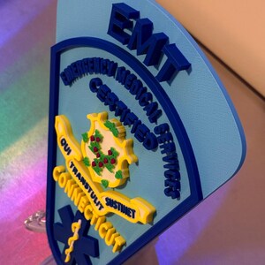Op de afbeelding: Een blauw en geel EMT-badge met de tekst "Connecticut" en "Certified Emergency Medical Services". De badge toont het staatszegel en een levensster-symbool.
