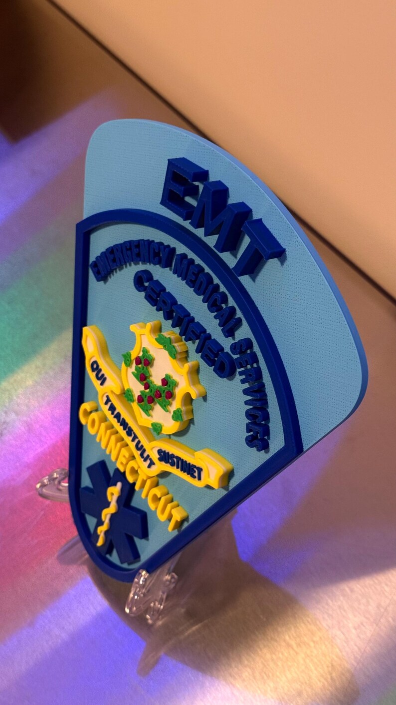 Op de afbeelding: Een blauw en geel EMT-badge met de tekst "Connecticut" en "Certified Emergency Medical Services". De badge toont het staatszegel en een levensster-symbool.