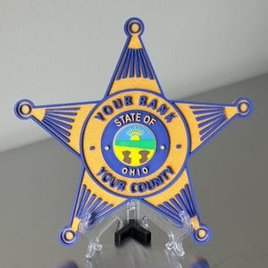 May include: A star-shaped badge with a gold-colored base and blue accents. The badge has the words "YOUR RANK", "STATE OF OHIO", and "YOUR COUNTY" printed on it. The center of the badge features a circular design with a landscape.