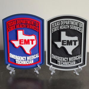 May include: Two shield-shaped wall plaques featuring the text Texas Department of State Health Services and Emergency Medical Technician with an EMT-labeled map of Texas. One plaque is blue and red, while the other is black and white.