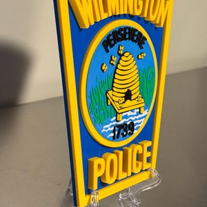 May include: A blue and yellow Wilmington Police Department sign. The sign features the words "Wilmington" and "Police" in yellow, with a blue center containing a beehive and the word "Persevere".