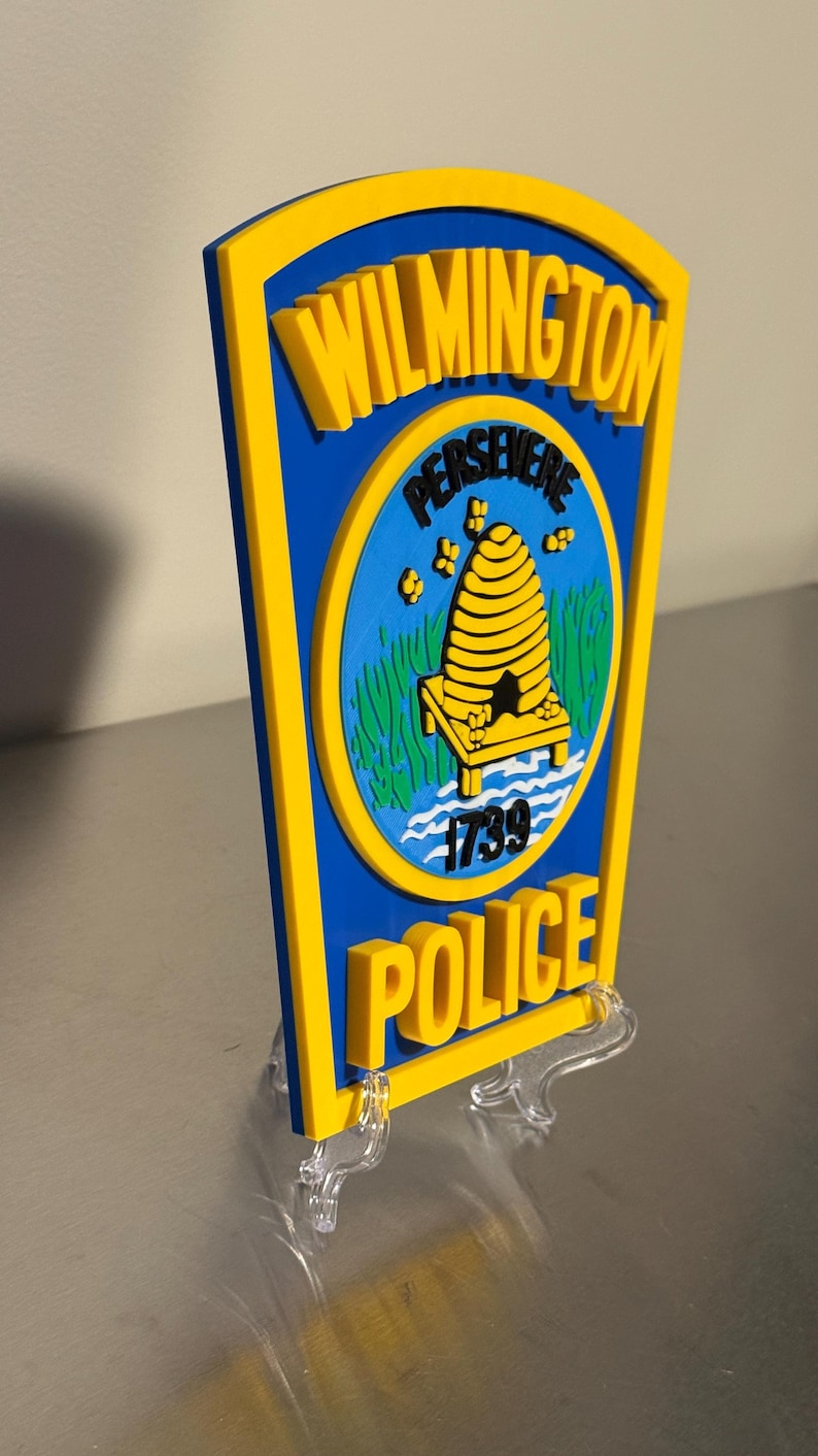 May include: A blue and yellow Wilmington Police Department sign. The sign features the words "Wilmington" and "Police" in yellow, with a blue center containing a beehive and the word "Persevere".