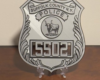 May include: A silver-toned shield-shaped plaque featuring the text SUFFOLK COUNTY N.Y. POLICE, a central circular emblem with an illustration of a bull, and the number 5502 displayed in a rectangular frame at the bottom with decorative scrollwork.