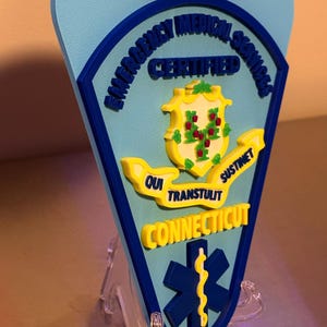 Op de afbeelding: Een lichtblauw en donkerblauw EMT-badge met de woorden "Emergency Medical Services Certified" en "Connecticut". De badge heeft een geel embleem met de woorden "Qui Transtulit Sustinet". Onderaan staat een blauw symbool van de levensster.
