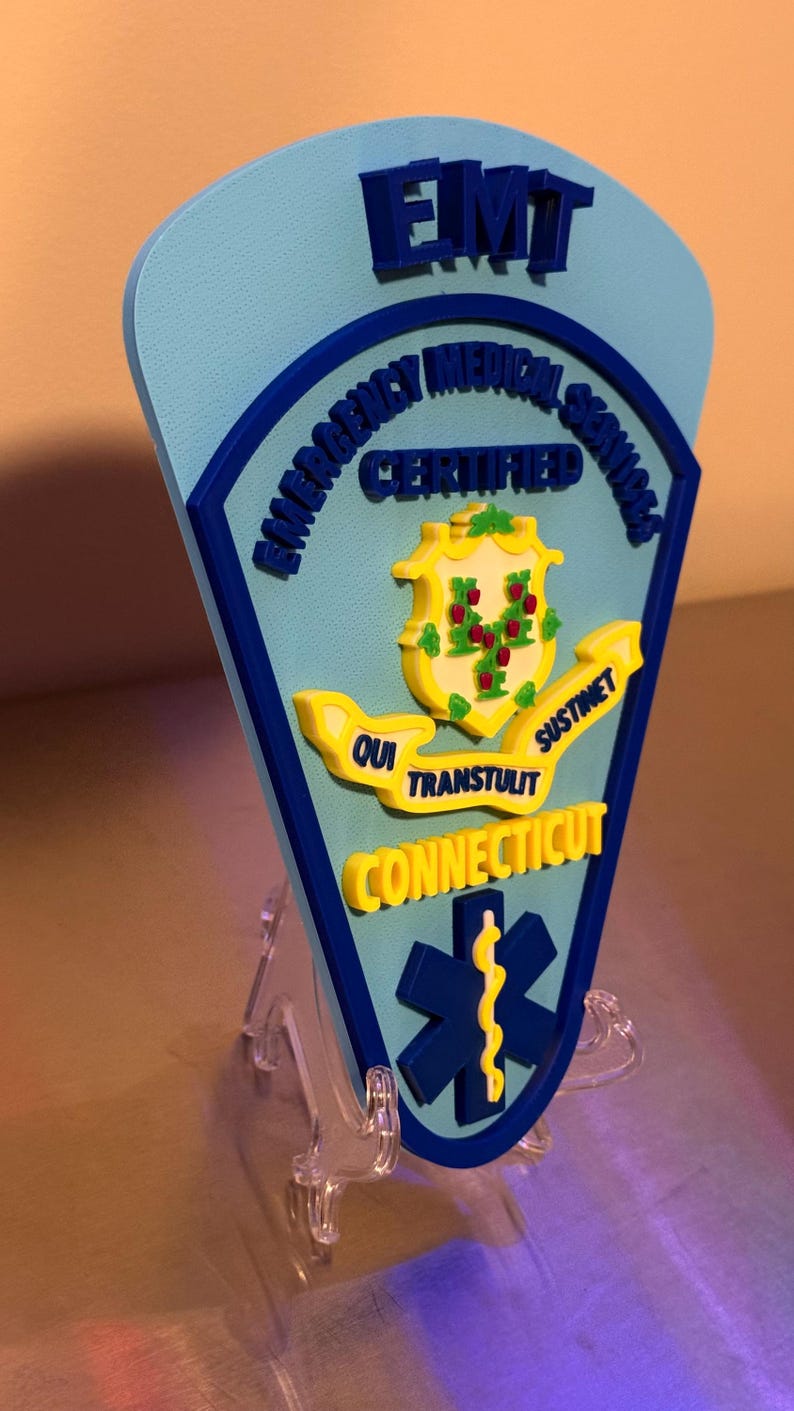 Op de afbeelding: Een lichtblauw en donkerblauw EMT-badge met de woorden "Emergency Medical Services Certified" en "Connecticut". De badge heeft een geel embleem met de woorden "Qui Transtulit Sustinet". Onderaan staat een blauw symbool van de levensster.