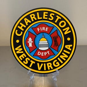 May include: A round emblem with the text "CHARLESTON WEST VIRGINIA" in yellow on a black and yellow border. The center features a fire department emblem with a fire hydrant, a building, and the words "FIRE DEPT".