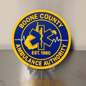 May include: A round sign with a blue background and yellow border. The sign reads "BOONE COUNTY AMBULANCE AUTHORITY" in yellow text. The center features a yellow star of life symbol and the text "EST. 1980".