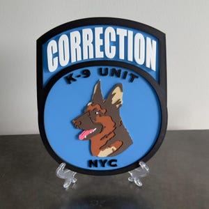 May include: A shield-shaped sign with the words "CORRECTION K-9 UNIT NYC" in white lettering. The sign features a blue background with a detailed illustration of a German Shepherd dog. The sign is black around the edges.