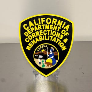 May include: A shield-shaped emblem featuring the text CALIFORNIA DEPARTMENT OF CORRECTIONS & REHABILITATION in yellow letters on a black background. The center displays a circular seal with a landscape, a figure, a bear, and the word EUREKA.