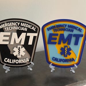 May include: Two EMT (Emergency Medical Technician) badges, one black and one blue, displayed on clear stands. Each badge features the text "Emergency Medical Technician," the letters "EMT," a medical symbol, and the word "California."