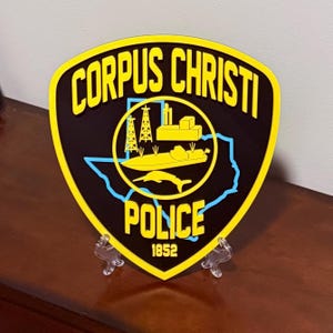 May include: A shield-shaped emblem with the words "CORPUS CHRISTI POLICE" in yellow lettering. The emblem features a black background with a yellow border and a blue outline of Texas. The year "1852" is at the bottom.