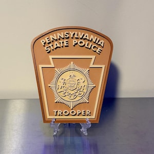May include: A tan and cream-colored Pennsylvania State Police Trooper plaque featuring a keystone shape with the text PENNSYLVANIA STATE POLICE at the top and TROOPER at the bottom, centered around a detailed circular emblem with a central crest.