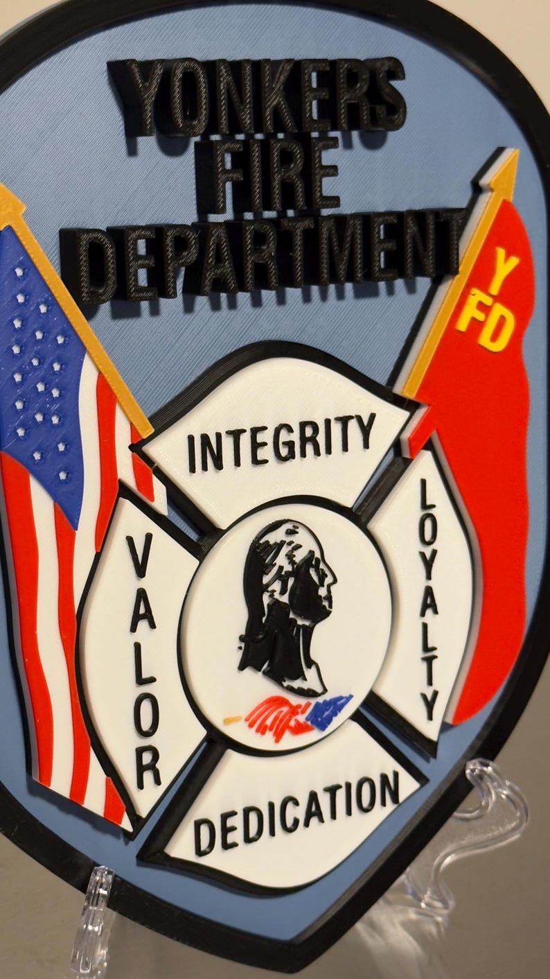 Op de afbeelding: Een 3D-geprint embleem voor de brandweer van Yonkers. Het ontwerp bevat de tekst "YONKERS FIRE DEPARTMENT", een Amerikaanse vlag, een rode vlag en de woorden "INTEGRITY", "VALOR", "LOYALTY" en "DEDICATION".
