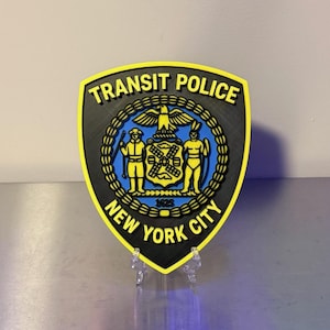 May include: A shield-shaped Transit Police New York City emblem featuring a black background with a bright yellow border. The center displays a blue circular seal with a golden eagle, two figures, and the year 1625, all rendered in yellow.