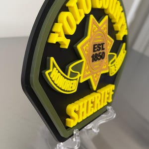Peut inclure: Un badge de sh&eacute;rif du comt&eacute; de Yolo imprim&eacute; en 3D, en noir, jaune et vert, pr&eacute;sent&eacute; sur un support transparent. Le badge comporte les mots "YOLO COUNTY SHERIFF" et "HONORED" avec un embl&egrave;me d'&eacute;toile et l'ann&eacute;e "EST 1850".