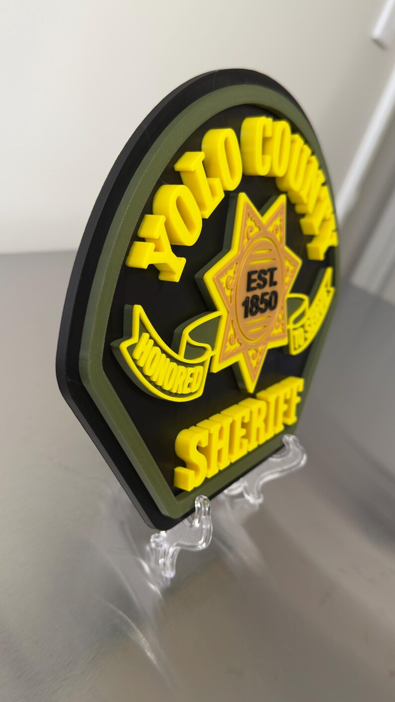 Peut inclure: Un badge de sh&eacute;rif du comt&eacute; de Yolo imprim&eacute; en 3D, en noir, jaune et vert, pr&eacute;sent&eacute; sur un support transparent. Le badge comporte les mots "YOLO COUNTY SHERIFF" et "HONORED" avec un embl&egrave;me d'&eacute;toile et l'ann&eacute;e "EST 1850".