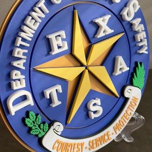 May include: A blue and gold round plaque with the words "Department of Public Safety" and "Texas" in white letters. The plaque features a five-pointed star with the letters "Texas" around it. The plaque also has the words "Courtesy - Service - Protection" in white letters on a scroll.