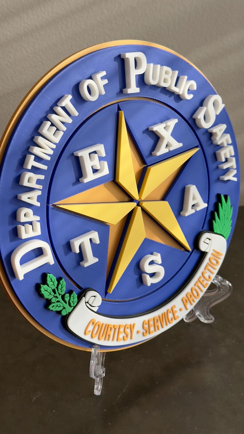 May include: A blue and gold round plaque with the words "Department of Public Safety" and "Texas" in white letters. The plaque features a five-pointed star with the letters "Texas" around it. The plaque also has the words "Courtesy - Service - Protection" in white letters on a scroll.