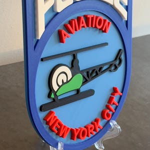 May include: A blue and white 3D printed sign with the words "POLICE AVIATION NEW YORK CITY" in red and white lettering. A black helicopter with a green rotor is in the center of the sign.