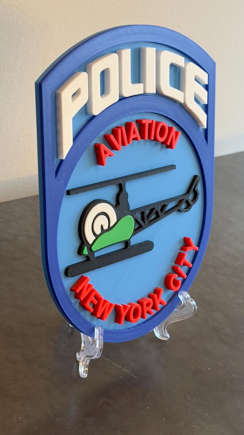 May include: A blue and white 3D printed sign with the words "POLICE AVIATION NEW YORK CITY" in red and white lettering. A black helicopter with a green rotor is in the center of the sign.