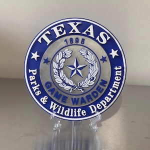 May include: A round, blue and silver Texas Game Warden Department sign. The sign features the text "TEXAS 1895" at the top, with a star and wreath design in the center. The words "Parks & Wildlife Department" are around the edge.