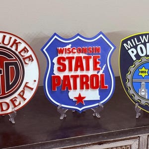 May include: Three decorative signs: Milwaukee Fire Dept. in red, black, and white; Wisconsin State Patrol in blue, white, and red; and Milwaukee Police in navy, yellow, and blue. Each sign is displayed on a clear stand.