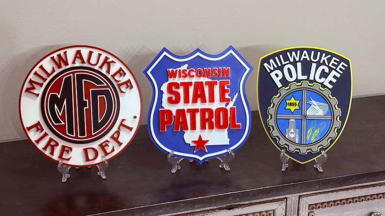 May include: Three decorative signs: Milwaukee Fire Dept. in red, black, and white; Wisconsin State Patrol in blue, white, and red; and Milwaukee Police in navy, yellow, and blue. Each sign is displayed on a clear stand.