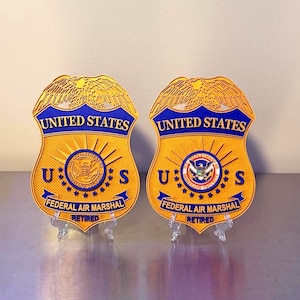 May include: Two gold and blue Federal Air Marshal badges with the words "UNITED STATES" across the top. The badges have a shield shape with the text "FEDERAL AIR MARSHAL" and "RETIRED" at the bottom.