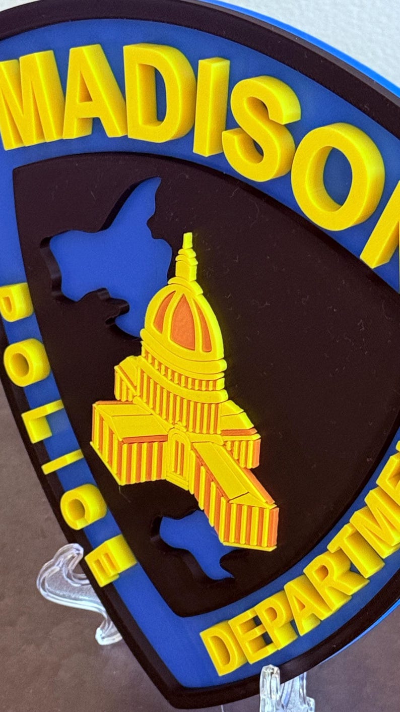 May include: A badge with the words "MADISON POLICE DEPARTMENT" in yellow, and a yellow and orange building design on a black background, set against a blue backdrop. The badge is shield-shaped.
