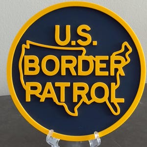 May include: A circular sign featuring a dark blue background with a yellow border. The text U.S. BORDER PATROL is displayed in raised yellow lettering, with the outline of the United States map integrated behind the word BORDER PATROL.