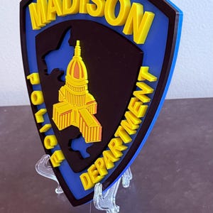 May include: A shield-shaped emblem for the Madison Police Department. The emblem features the words "MADISON POLICE DEPARTMENT" in yellow, with a yellow building in the centre. The background is black and blue.