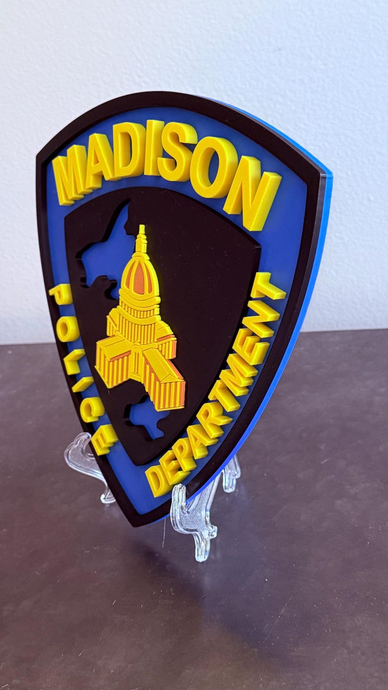 May include: A shield-shaped emblem for the Madison Police Department. The emblem features the words "MADISON POLICE DEPARTMENT" in yellow, with a yellow building in the centre. The background is black and blue.