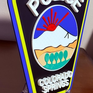 K&ouml;nnte beinhalten: Ein 3D-gedrucktes Polizeibadge mit blauem Hintergrund, einem wei&szlig;en Berg mit einer roten Sonne und gr&uuml;nen B&auml;umen. Das Abzeichen zeigt "POLICE" in wei&szlig;en Buchstaben und "COLORADO SPRINGS" in wei&szlig;en Buchstaben unten.